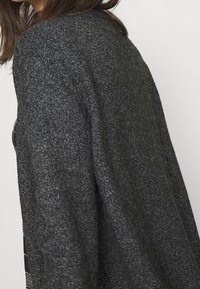 Černý pletený kardigan s texturou, který má volný střih a široké rukávy. Tkanina se zdá být měkká s jemným leskem, což zvyšuje celkový design.