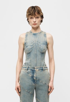 Γυναίκα με κοντά μαλλιά, ντυμένη με αμάνικο στενό τοπ από ξεβαμμένο τζιν και ψηλόμεσο παντελόνι από τζιν που ταιριάζει, στέκεται μπροστά σε απλό φόντο.