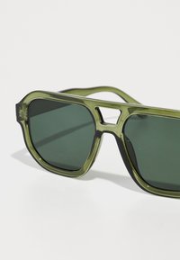 Ochelari de soare cu tentă verde, cu un cadru gros, transparent, de culoare verde oliva. Lentile pătrate și un detaliu distinctiv sub formă de decupaj pe punte.