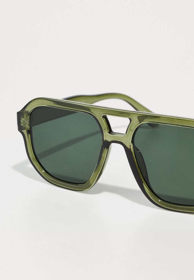 Lunettes de soleil teintées de vert avec une monture épaisse et transparente olive. Verres carrés et un détail découpé distinctif sur le pont.