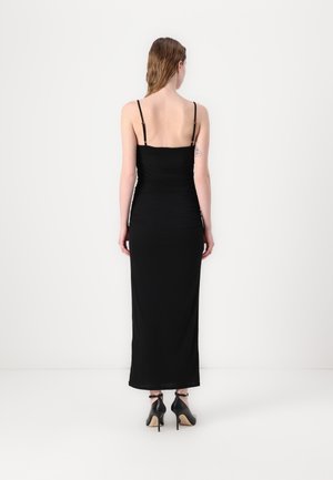 Femme portant une robe longue noire sans manches avec de fines bretelles et des chaussures à talons hauts noires, debout dos à un mur uni.