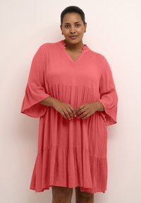 Vestido rosa coral hecho de una tela ligera, con escote en V, mangas acampanadas y un diseño en capas. Textura suave con un ajuste holgado.