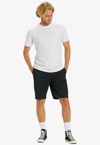 T-shirt bianca a girocollo abbinata a pantaloni corti scuri. Il modello sta in piedi con le mani in tasca, indossando sneakers alte nere e calzini bianchi.
