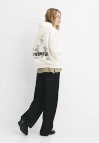 Vit huva sweatshirt med grå stjärna och textgrafik på ryggen; bärs över en ljus skjorta, i kombination med lösa svarta byxor och svarta skor.
