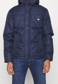 Marinblå vadderad jacka med quiltad textur, dragkedja fram och justerbar huva. Har logotypdetalj på vänster bröst och sidofickor.
