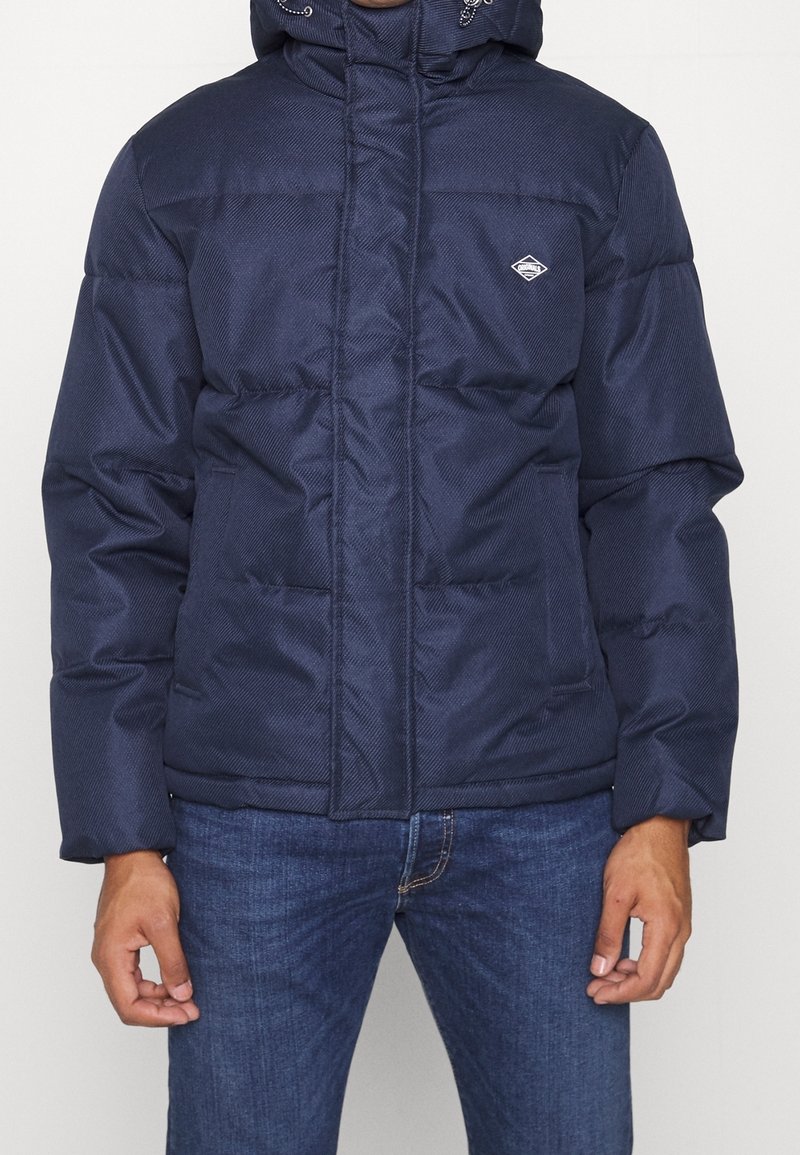 Marinblå vadderad jacka med quiltad textur, dragkedja fram och justerbar huva. Har logotypdetalj på vänster bröst och sidofickor.