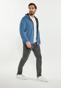 Felpa blu con zip e dettagli gialli sopra una maglietta bianca, abbinata a jeans grigi e sneakers bianche. Tessuto morbido e design aderente.