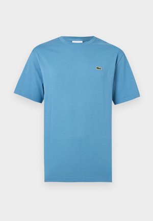 T-shirt bleu à manches courtes avec col rond et petit logo de crocodile vert sur le côté gauche de la poitrine, présenté sur fond blanc.