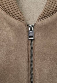 Bruine ritsjack met een geribde kraag, gladde stof en een metalen rits met een logodetail, die de texturen en materialen benadrukt.