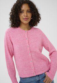 Pink knitted cardigan with a round neck, silver button closures, and ribbed cuffs. Soft texture with a relaxed fit, paired with blue jeans.