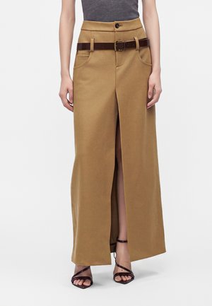 Femme portant un pantalon camel taille haute à jambes larges avec une fente à l'avant, ceinture marron, top gris et talons noirs à brides sur fond blanc.