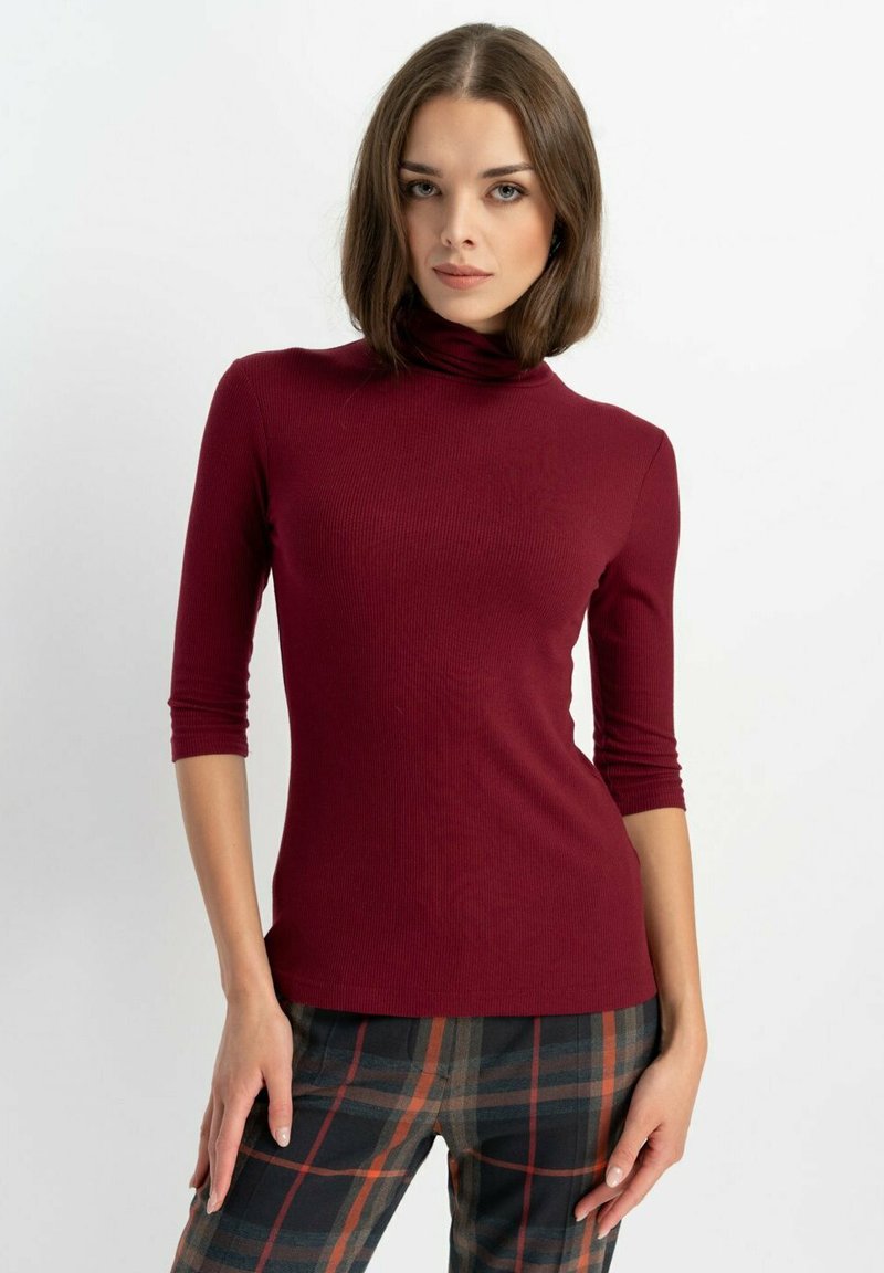 Burgundowy golf prążkowany z rękawami do trzech czwartych, w połączeniu z spodniami w kratę z pomarańczowymi i zielonymi akcentami na neutralnym tle.