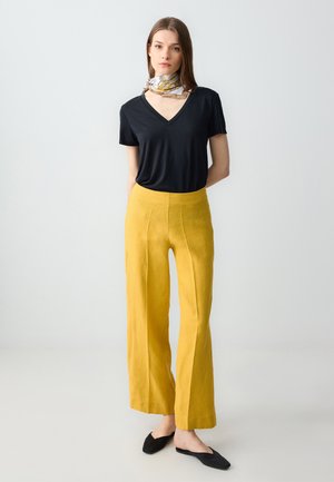 T-shirt nera a V, pantaloni gialli a gamba larga, texture del tessuto liscia, colori uni, abbinati a scarpe nere slip-on e a una sciarpa a fantasia.