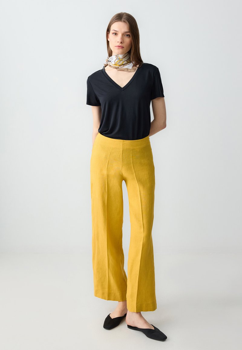 Zwarte V-hals T-shirt, gele wijde broek, gladde stoffen textuur, effen kleuren, gecombineerd met zwarte instapschoenen en een gestreepte sjaal.