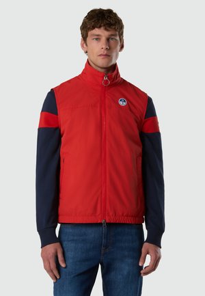 Joven con un chaleco rojo sin mangas con cremallera sobre una camisa azul marino de manga larga con rayas rojas en las mangas, de pie contra un fondo liso.