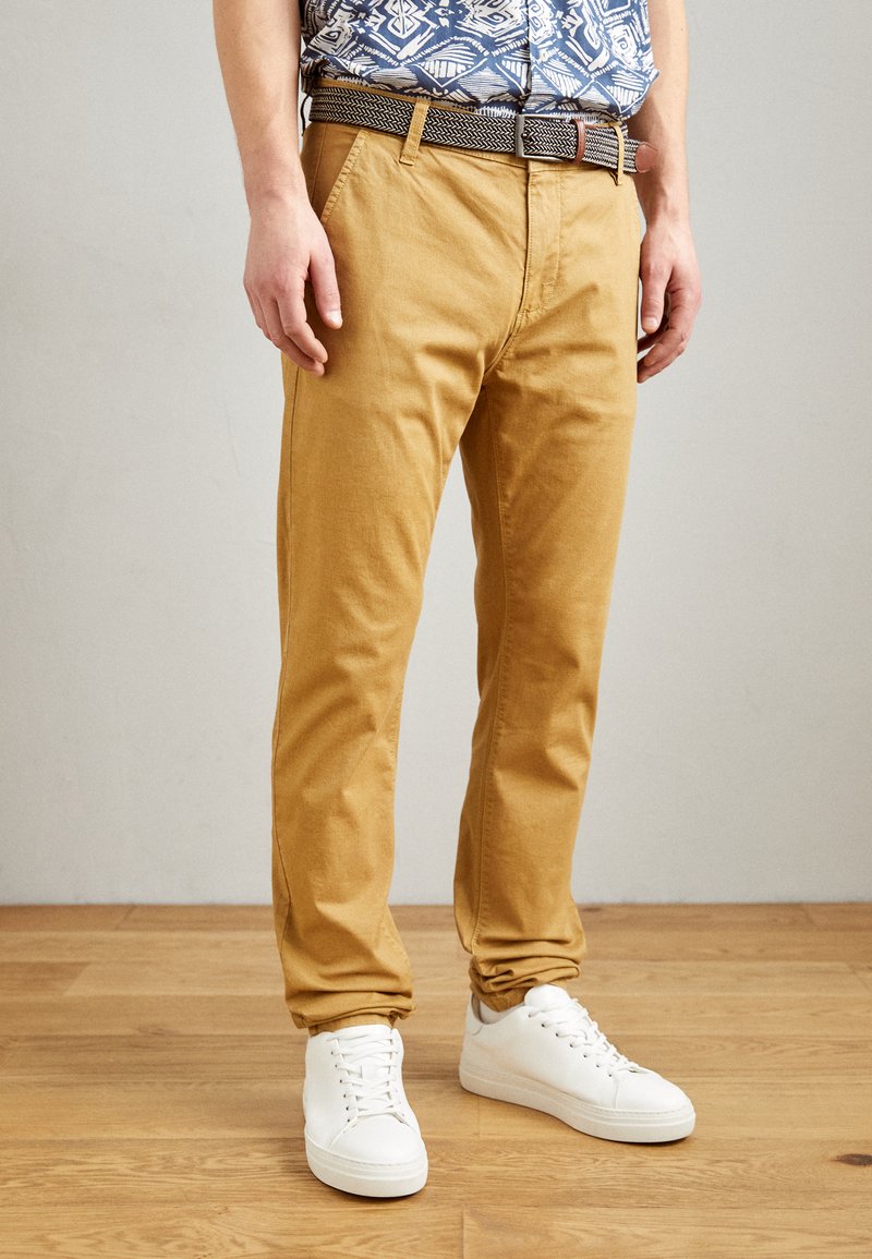 Chino bej cu croi drept, din material de bumbac, dotat cu o curea texturată țesută și asociat cu adidași albi. Finisaj neted, stil casual.