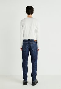 Jeans de mezclilla de color azul oscuro con un corte recto, que presentan bolsillos traseros y una etiqueta de piel en la cinturilla. Llevados con una camisa blanca de manga larga.