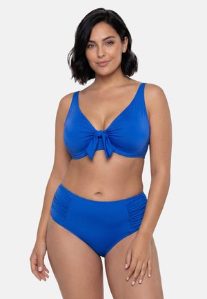 Kvinde med kort sort hår iført blå bikini med knyttet top og højtaljede underdele, stående mod en ensfarvet lys baggrund.