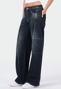Lange, donkere spijkerbroek met een hoge taille, subtiele vervaging, grote zijzakken en contrasterende stiksels. Gecombineerd met witte sneakers.