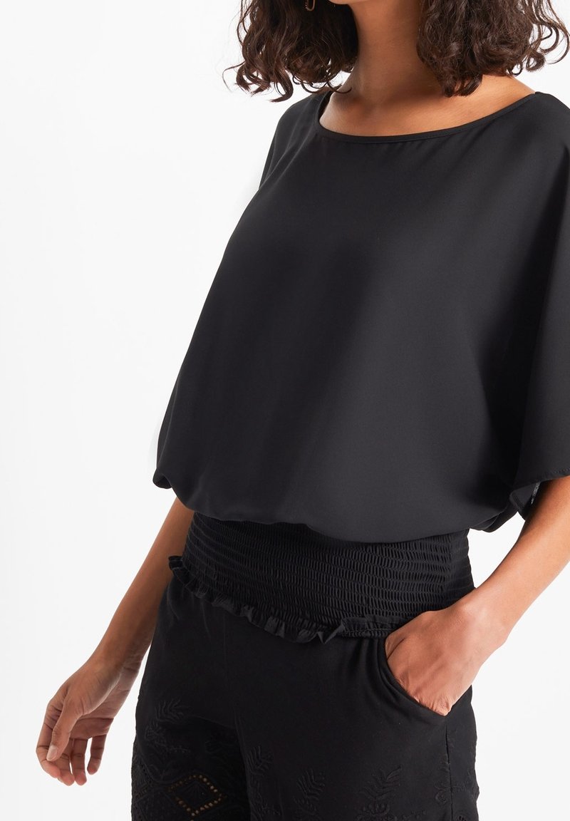 Femme portant un haut noir ample à larges manches courtes et un pantalon noir taille haute avec une ceinture froncée, main dans la poche.