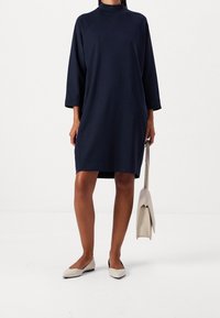 Mekko on laivastonsininen, ylisuuri ja pehmeää kangasta, jossa on korkea kaula-aukko ja kolmannes hihasta. Yhdistetty vaaleanbeigeihin litteisiin kenkiin ja kermanväriseen laukkuun.