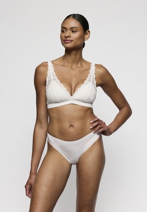 Vrouw die een witte kanten beha en bijpassend wit bikini-ondergoed draagt, staand met één hand op haar heup tegen een effen achtergrond.