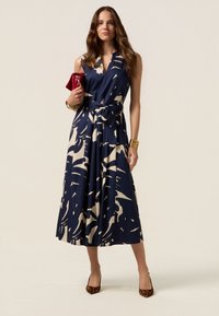 Navy blue sleeveless dress with a cream abstract pattern, a defined waist, and a flowy skirt. Accessories include a red clutch and gold bracelet.