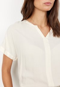 Lys beige bluse med korte ærmer, V-udskæring og subtil glans. Har en central søm og en glat, flydende stof tekstur.