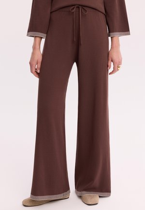 Brązowe spodnie o szerokich nogawkach z paskowanymi mankietami, talią ze sznurkiem, noszone z beżowymi teksturowanymi płaskimi butami i złotym pierścieniem na prawej ręce.