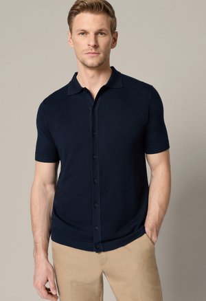 Młody mężczyzna o krótkich blond włosach, ubrany w granatową, krótkim rękawem koszulę zapinaną na guziki oraz beżowe spodnie, stojący na neutralnym tle.