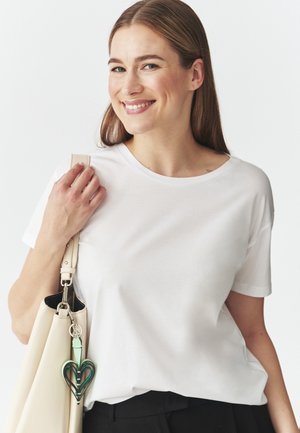 T-shirt bianco a maniche corte con scollatura rotonda, abbinato a una borsa crema con un portachiavi a forma di cuore verde e nero.