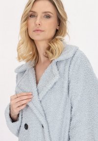 Jasnoniebieski, puszysty płaszcz o podwójnym zapięciu, z ząbkowanym kołnierzem i dwoma czarnymi guzikami. Miękka tekstura, odpowiednia na chłodną pogodę.
