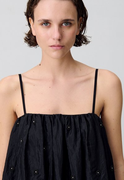 Vestido negro sin mangas hecho de una tela texturizada, que presenta detalles fruncidos y adornos circulares decorativos en todo su diseño.