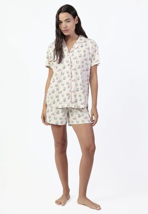Mujer joven descalza, vestida con pijama de manga corta abotonado y pantalones cortos a juego con estampado de animales de dibujos animados sobre un fondo claro.