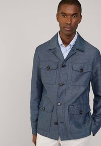 Niebieska tkaninowa kurtka z kołnierzem, czterema przednimi kieszeniami i czarnymi guzikami. Nałożona na białą koszulę w niebieskie paski. Gładka tekstura.