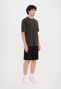 Cămășă neagră cu mâneci scurte, având un model geometric în nuanță de bej, combinată cu pantaloni scurți negri și adidași albi, creând o ținută casual.