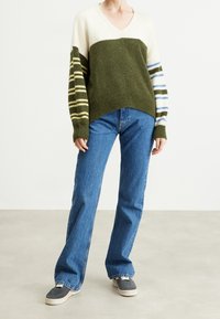 Sweter z dzianiny z dekoltem w kształcie litery V w kolorze oliwkowym i kremowym, z blokowym designem, z paskami na rękawach w kolorze oliwkowym, żółtym i niebieskim. Połączony z niebieskimi dżinsami.