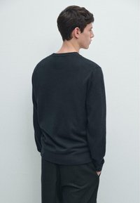 Maglione scuro in tessuto a maglia con scollo rotondo e polsini a costine, caratterizzato da una vestibilità morbida e colore uniforme, indossato con pantaloni scuri.
