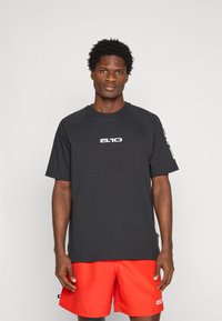 Čierne tričko s krátkym rukávom s bielym logom na hrudi, doplnené červenými šortkami s bielym značkovým akcentom. Klasický atletický strih.