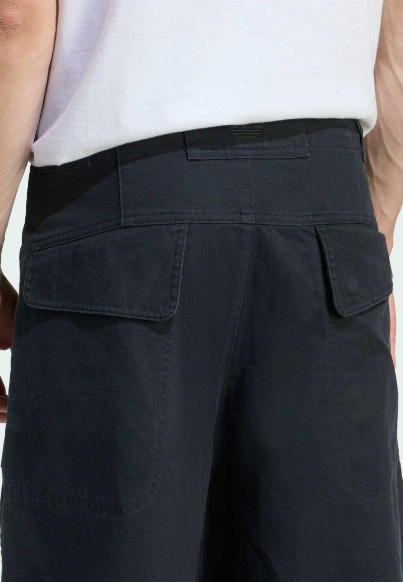 Man in zwarte broek met flap achterzakken en een wit shirt ingestopt, weergegeven vanaf de taille naar beneden en van achteraan.