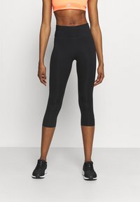 Atletisch persoon in zwarte high-waisted cropped leggings, zwarte sneakers en een oranje sportbeha, staande tegen een effen achtergrond.