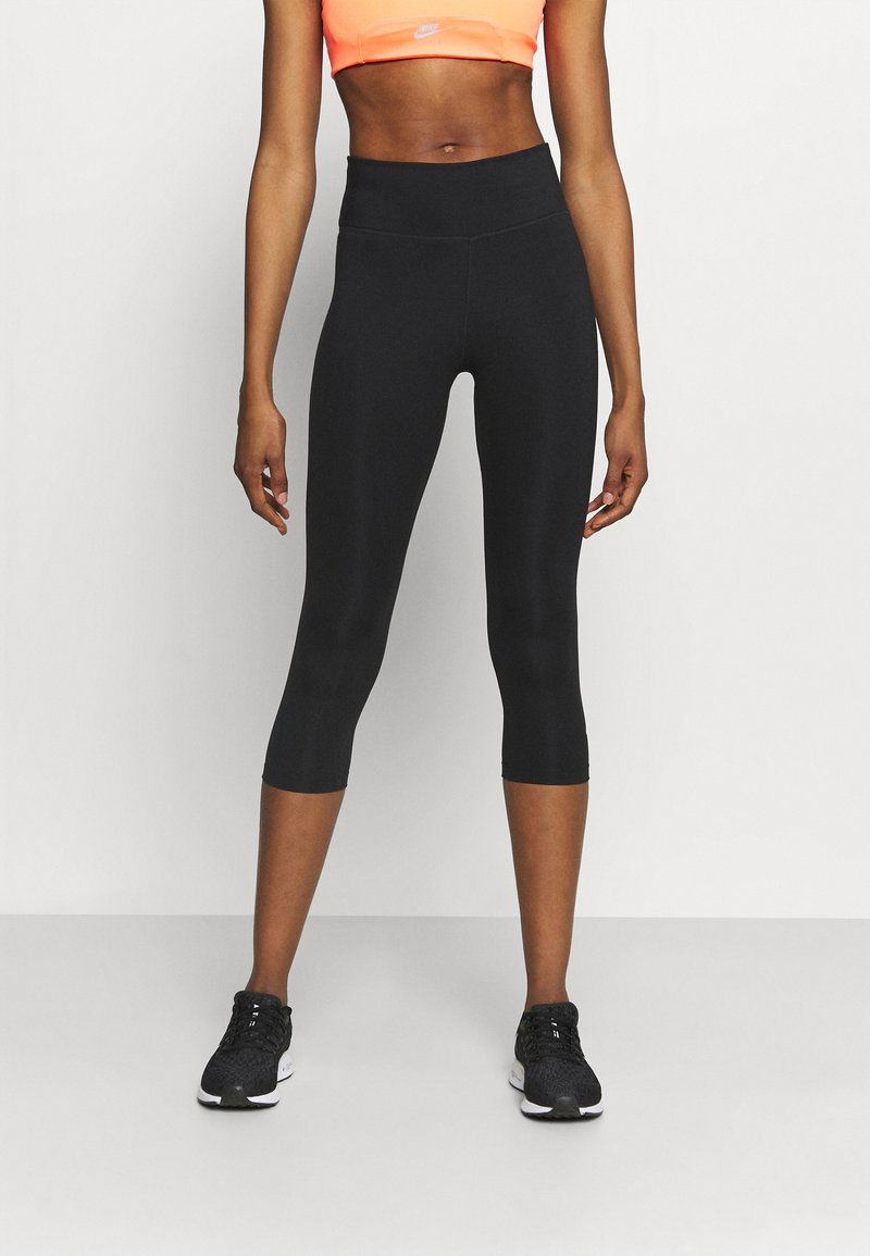 Atletisch persoon in zwarte high-waisted cropped leggings, zwarte sneakers en een oranje sportbeha, staande tegen een effen achtergrond.