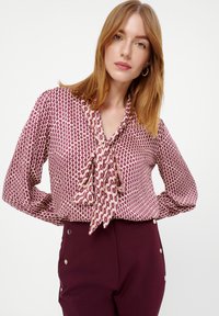 Langarm blouse in lichtcrème met een maroon geometrisch patroon, voorzien van een gestrikt decolleté en een zachte stof. Gecombineerd met maroon broek.
