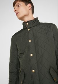 Quilted jacket in dark green with a high collar, gold buttons, and side pockets. Features a diagonal pattern and brown accents on the collar.