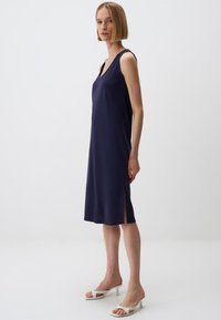 Granatowa sukienka bez rękawów z dekoltem w serek, gładką fakturą i bocznymi rozcięciami; modelka uzupełniła strój białymi sandałami na wysokim obcasie.