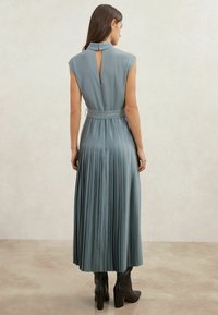 Plissé lange jurk in gedempt teal, met een hoge hals, mouwloze cap sleeves en een sleutelgat aan de achterkant, gecentreerd met een bijpassende riem. Gecombineerd met zwarte laarzen.