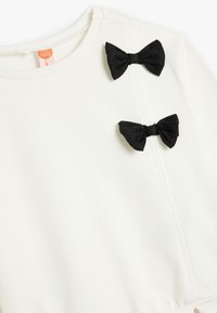 Sudadera blanca con lazos de tela negra cosidos en la parte delantera. El material parece suave, con un cuello acanalado y sutiles detalles de costura.