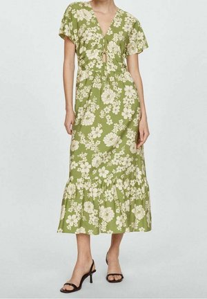 Femme portant une robe verte mi-longue à imprimé floral blanc, manches courtes, col en V avec un détail de nouage, et sandales noires à talons ouverts à l’avant.