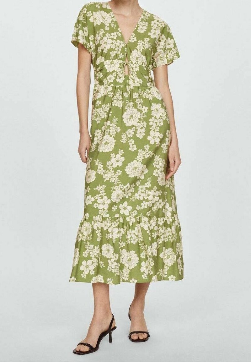 Femme portant une robe verte mi-longue à imprimé floral blanc, manches courtes, col en V avec un détail de nouage, et sandales noires à talons ouverts à l’avant.