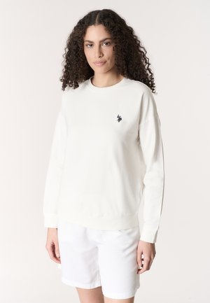 Donna con capelli ricci che indossa una felpa bianca a maniche lunghe con piccolo logo nero e pantaloncini bianchi, in piedi davanti a uno sfondo neutro.